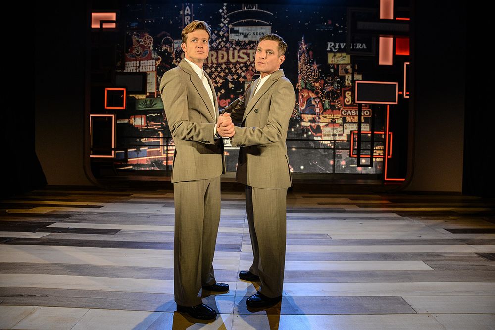 Two performers in beige suits stand together on a patterned floor with cityscape backdrop.