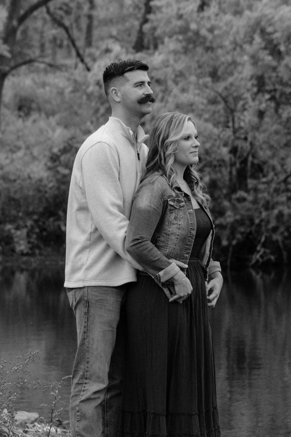 A black and white portrait of a couple standing by a lake in autumn.