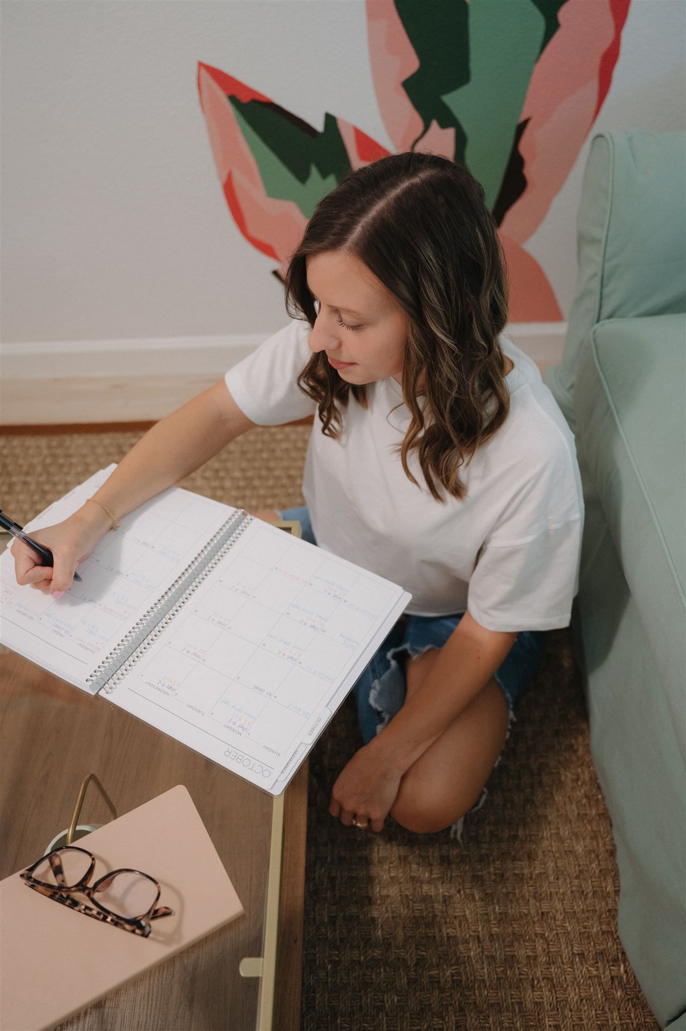 Someone writing in a notebook while sitting on a brown rug with pink wall art and a mint green couch in background.