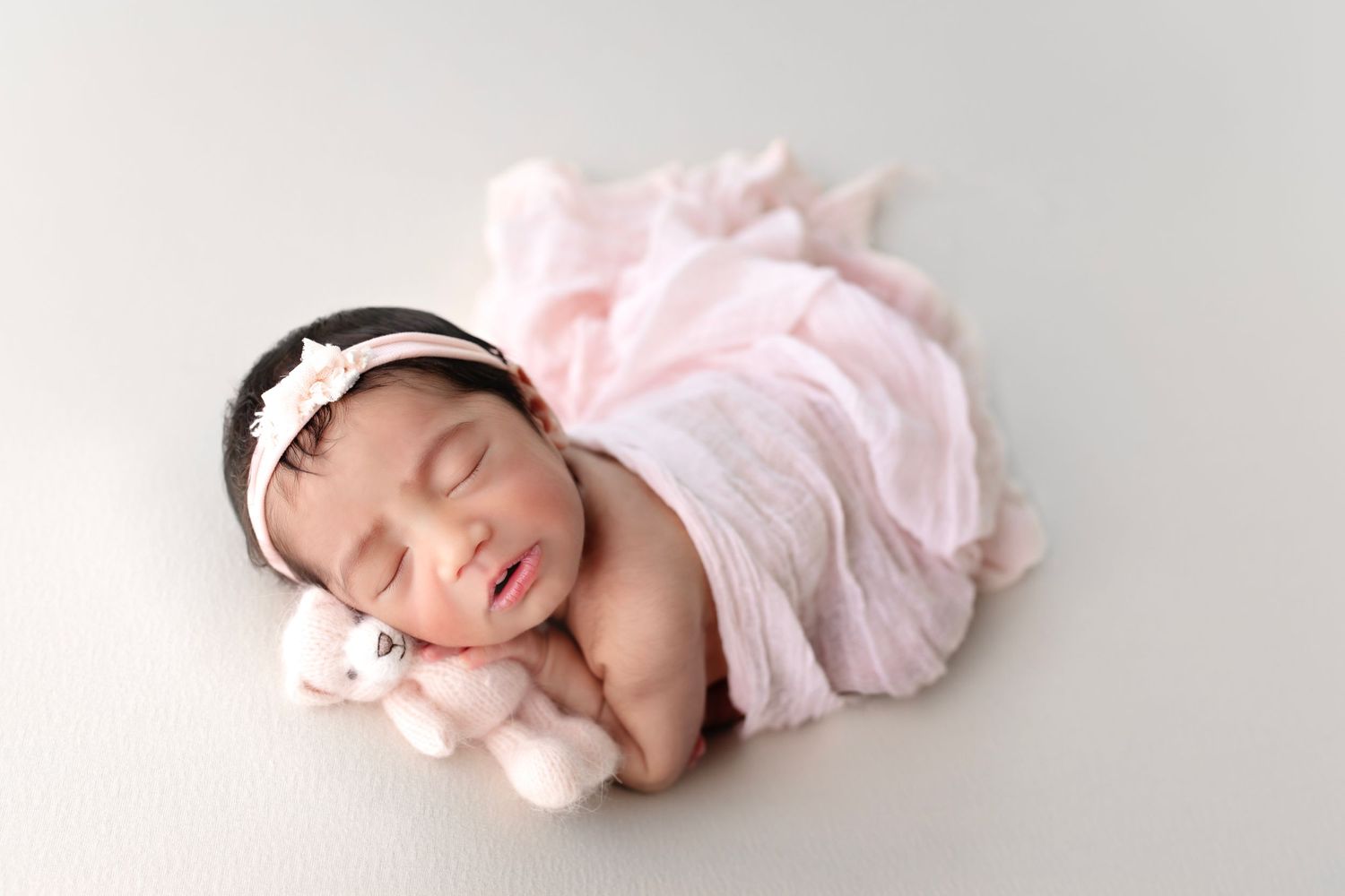 Sleeping newborn in pink wrap with headband and small plush toy on light background.