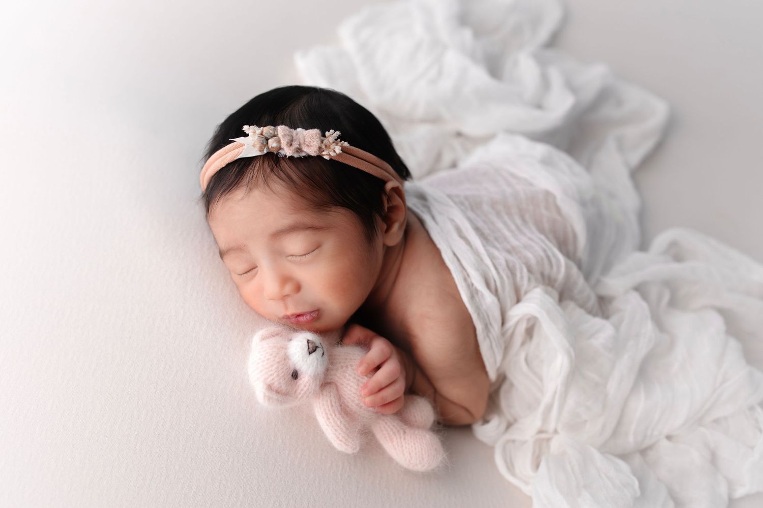 Peaceful newborn sleeping with white wrap and headband holding tiny plush toy.