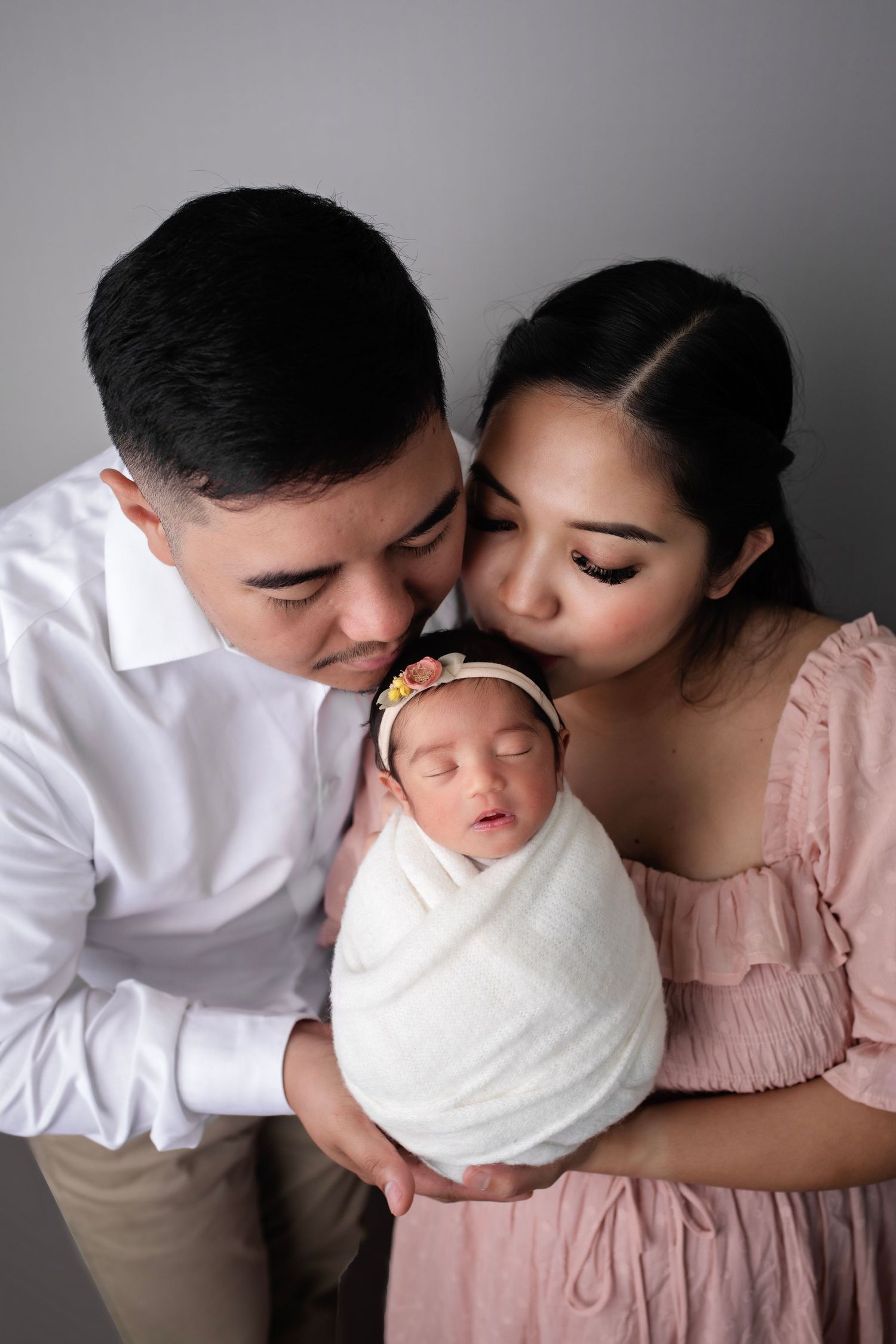 Parents in pink and white outfits share tender moment with swaddled newborn.