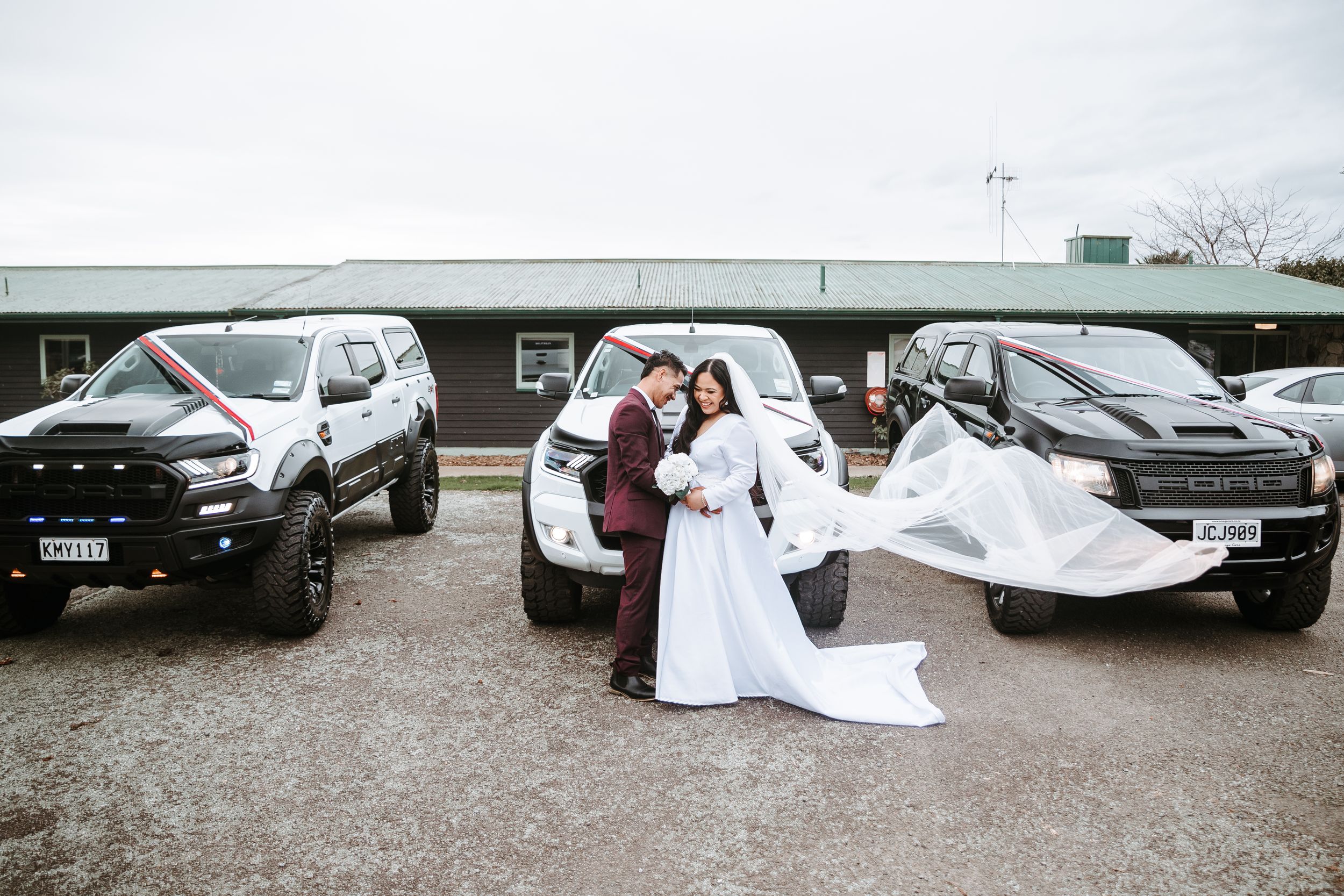 Bride and groom laughing in front of three wedding trucks in Manawatu - candid, relaxed New Zealand wedding photography.