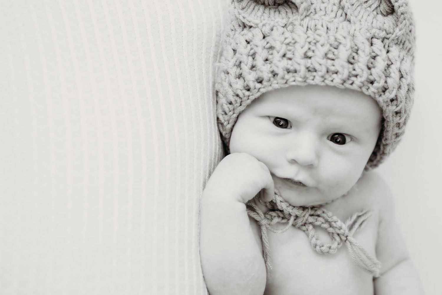 Baby wearing knit hat with hand near face in black and white portrait.