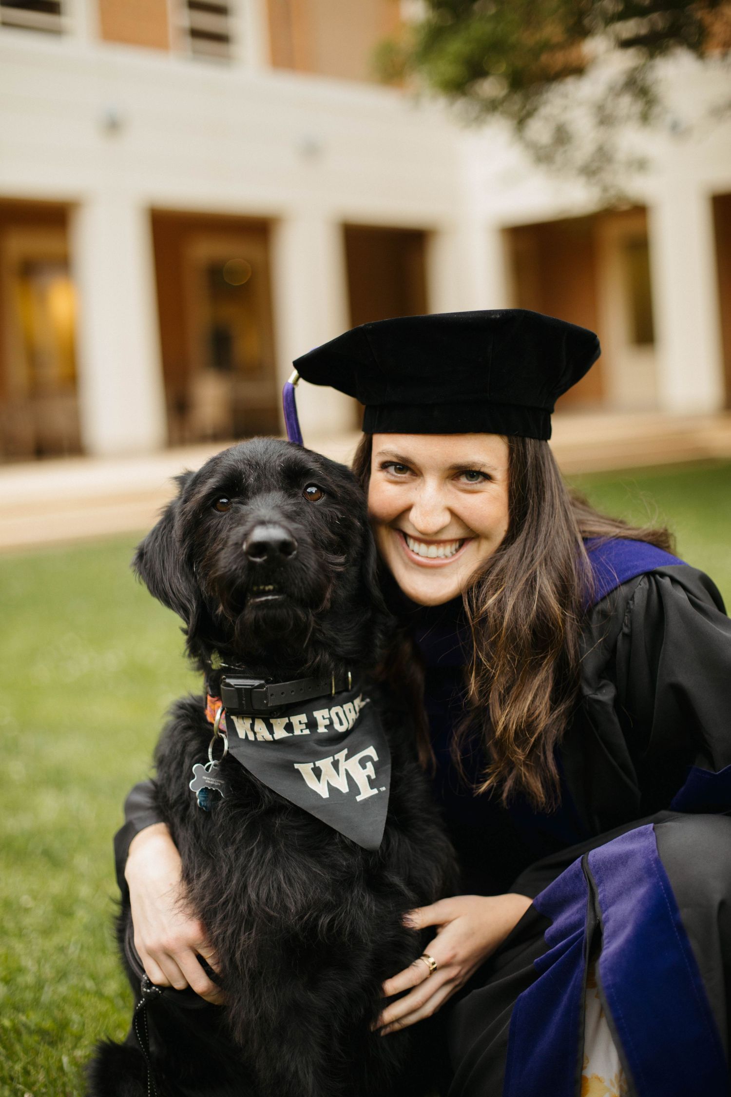Graduate in academic regalia poses with black dog wearing Wake Forest bandana on campus lawn.