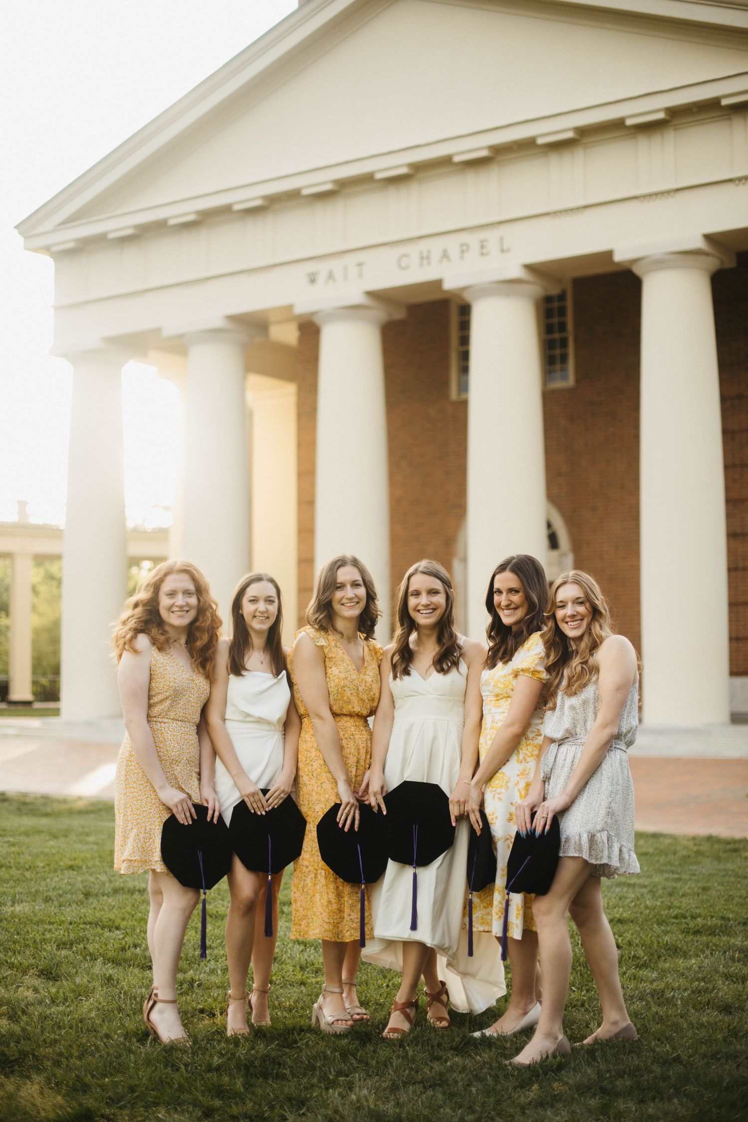 A group of friends in yellow and white dresses pose together in front of a chapel with white columns.