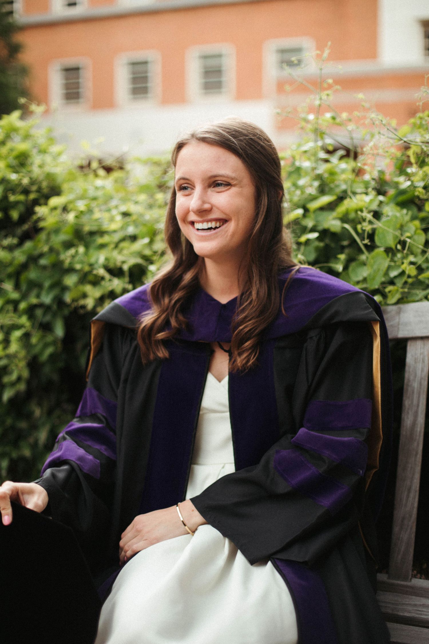 Graduate in academic regalia with purple and black gown sitting on bench in garden setting.