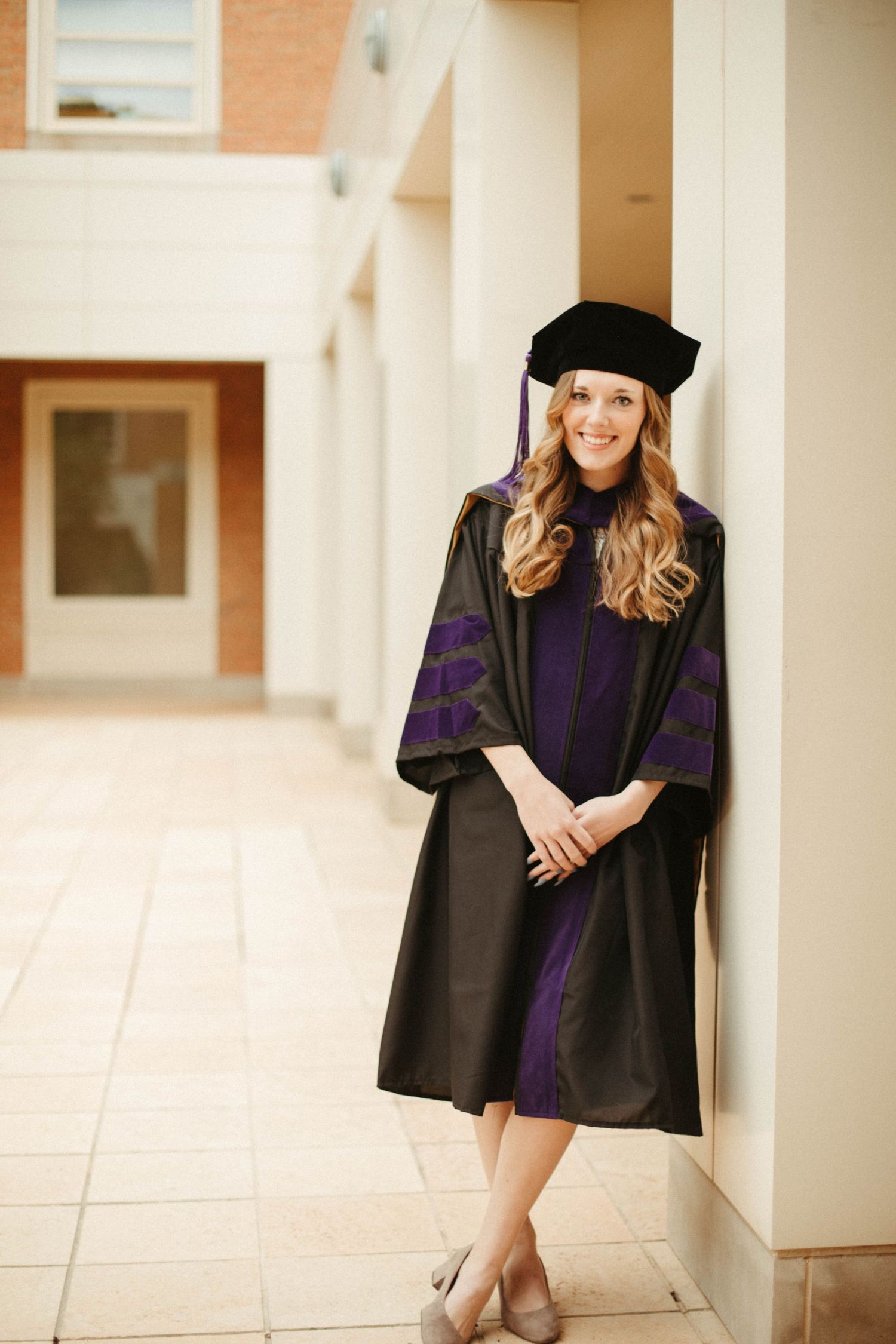 Graduate in purple and black academic regalia poses against white wall in hallway.