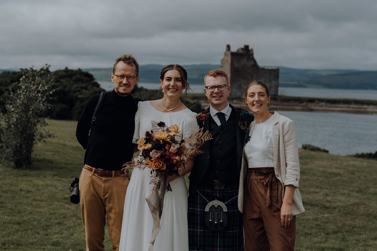 A wedding group photo against a dramatic Scottish landscape with a castle ruin in the distance.