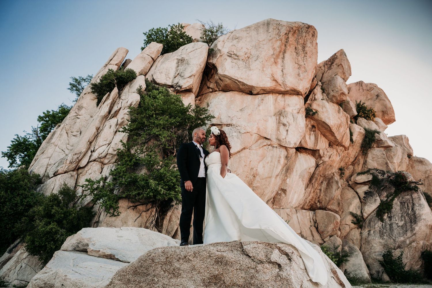 A couple in wedding attire embraces while standing on large granite boulders against a blue sky.