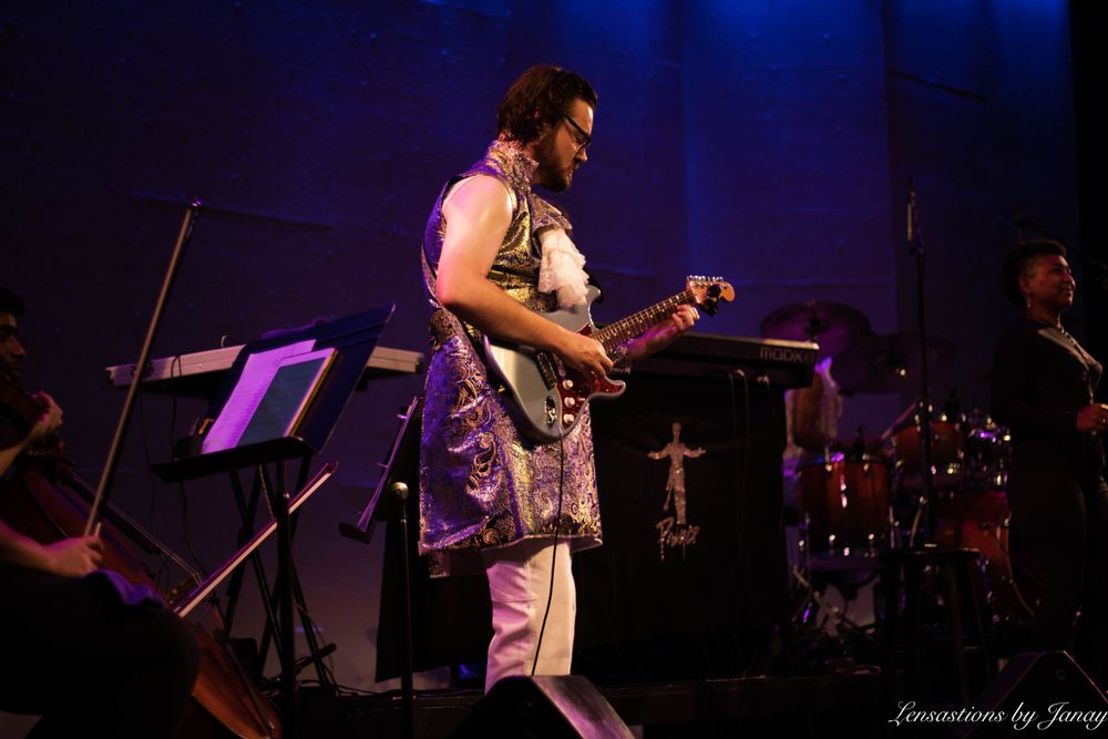 Guitarist performing on stage in a decorative vest under blue and purple stage lighting.