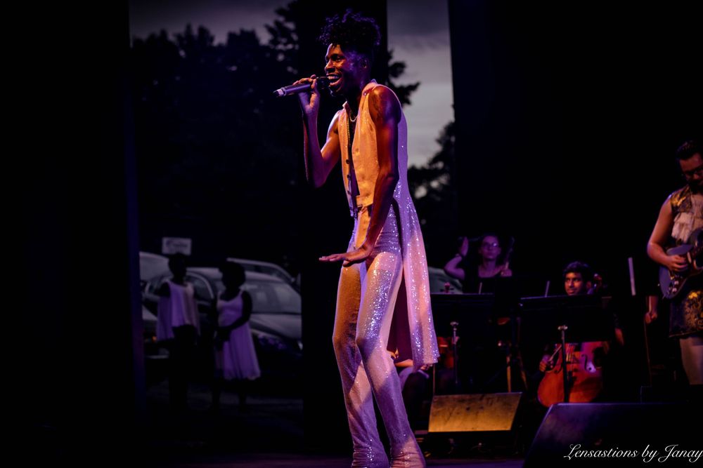 Performer singing on stage in a sparkly outfit under purple stage lights outdoors.