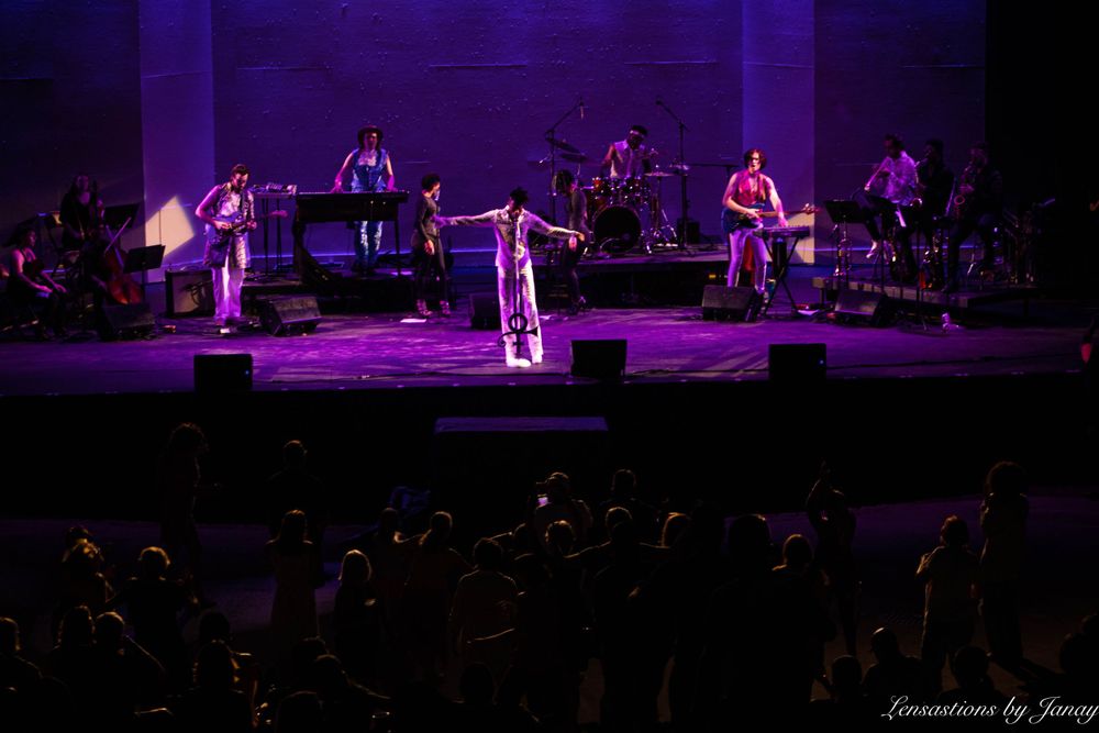 Female performer on stage with arms outstretched under purple lights before a live audience.