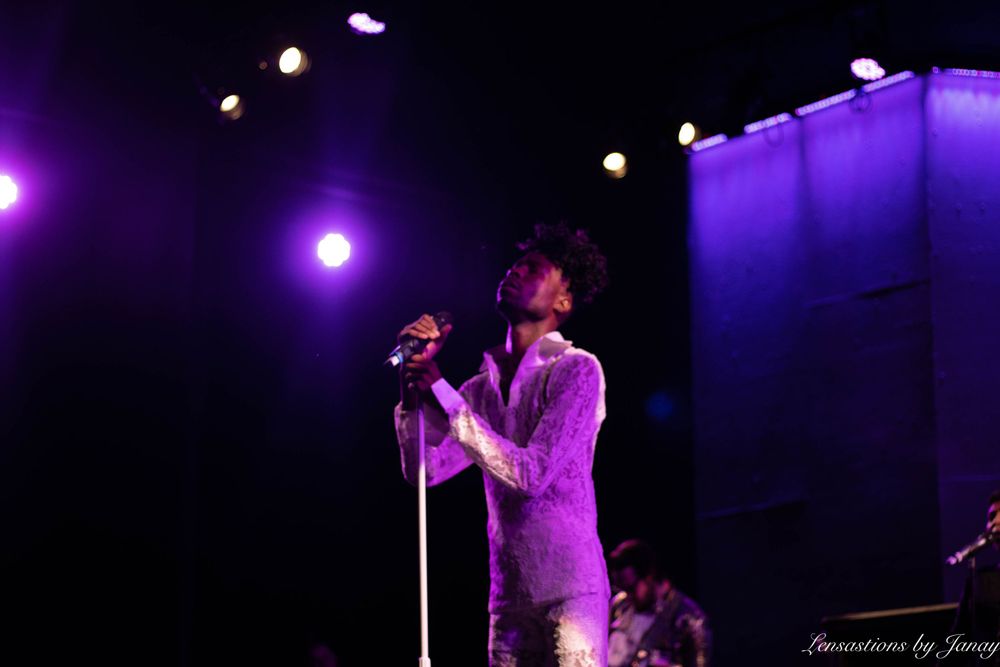 Singer performing on stage in a glittery outfit under dramatic purple and white lights.