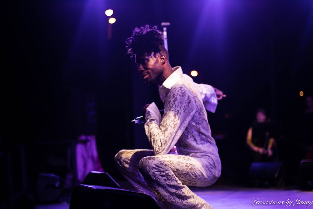 Male performer in white sequined outfit crouching on stage under purple concert lighting.