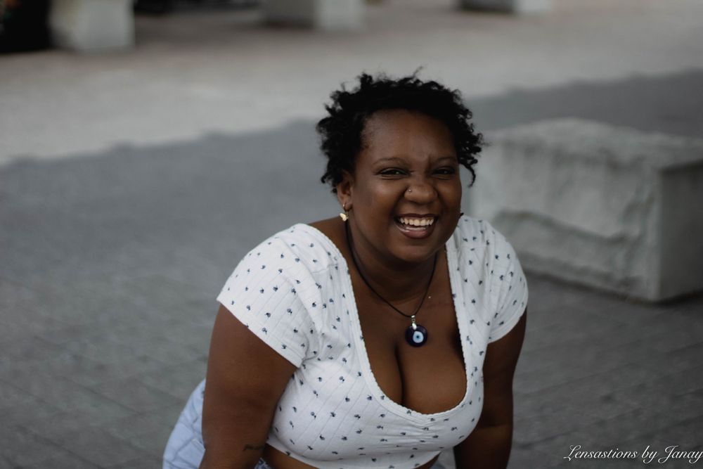 Smiling Black woman in white polka dot top sitting outdoors on a sunny day.