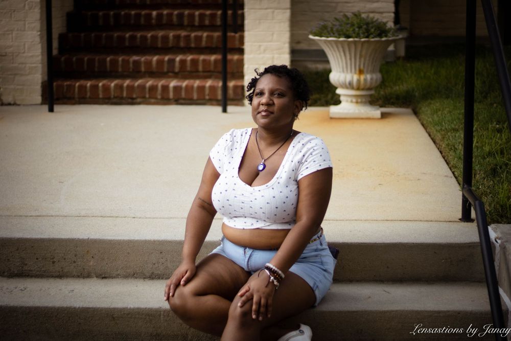 Young Black woman in white crop top and denim shorts sitting on outdoor steps smiling.