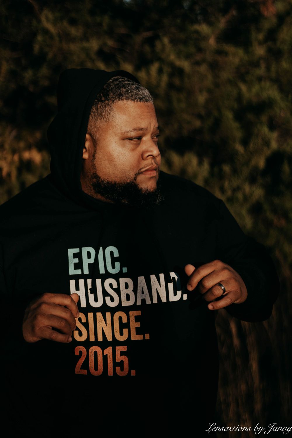 Man wearing an Epic Husband Since 2015 black hoodie posing outdoors near green foliage.