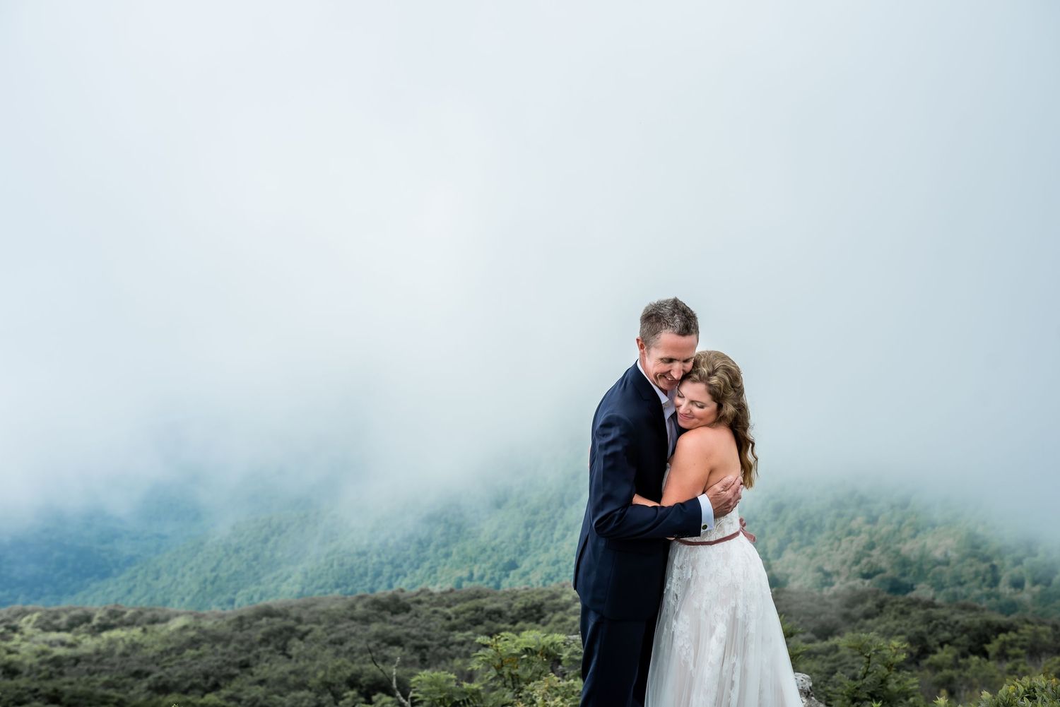 Man and woman embrace with mountains behind them and clouds surrounding them in Asheville, NC.