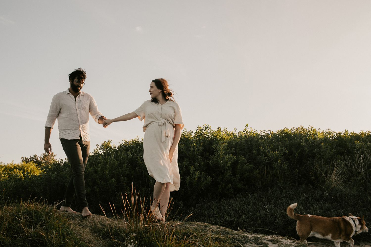 A pregnant couple holds hands while walking along a rocky path with their dog at golden hour.