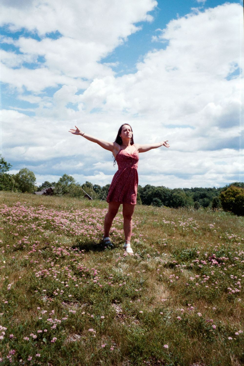 A person stands with arms wide open in a burgundy dress amid a field of scattered wildflowers.