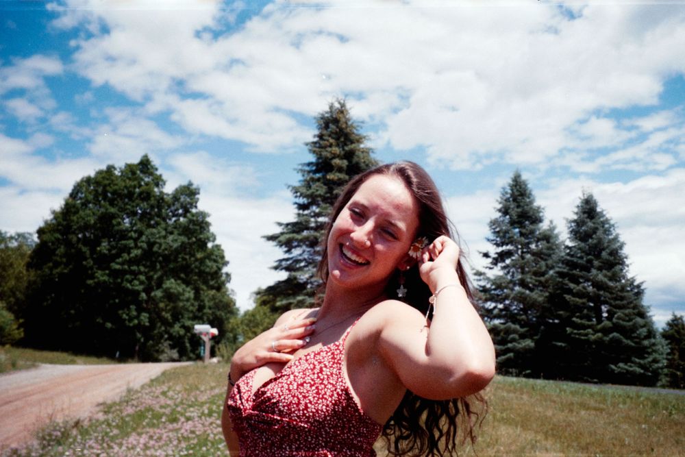 Someone in a floral dress poses cheerfully against a backdrop of evergreen trees on a sunny day.