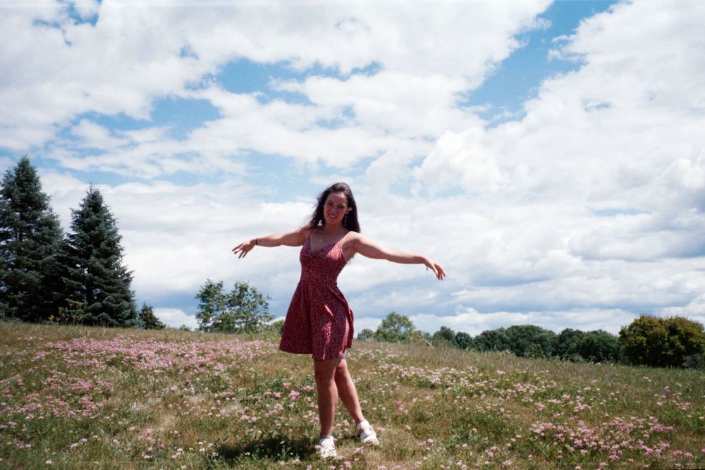 A person in a burgundy dress stands with arms outstretched in a wildflower meadow on a sunny day.