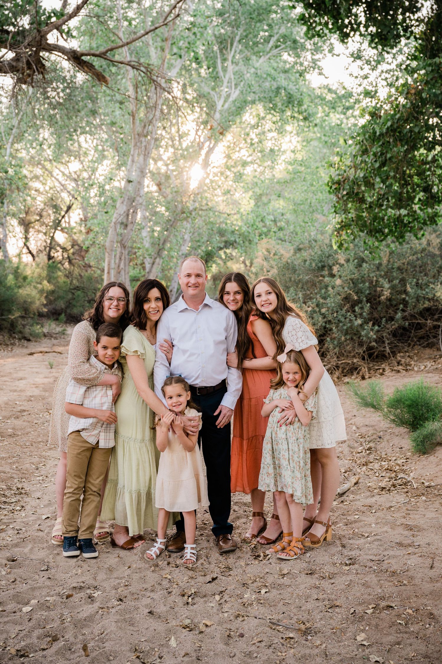A family dressed in earth-toned outfits poses together on a sunlit nature trail.