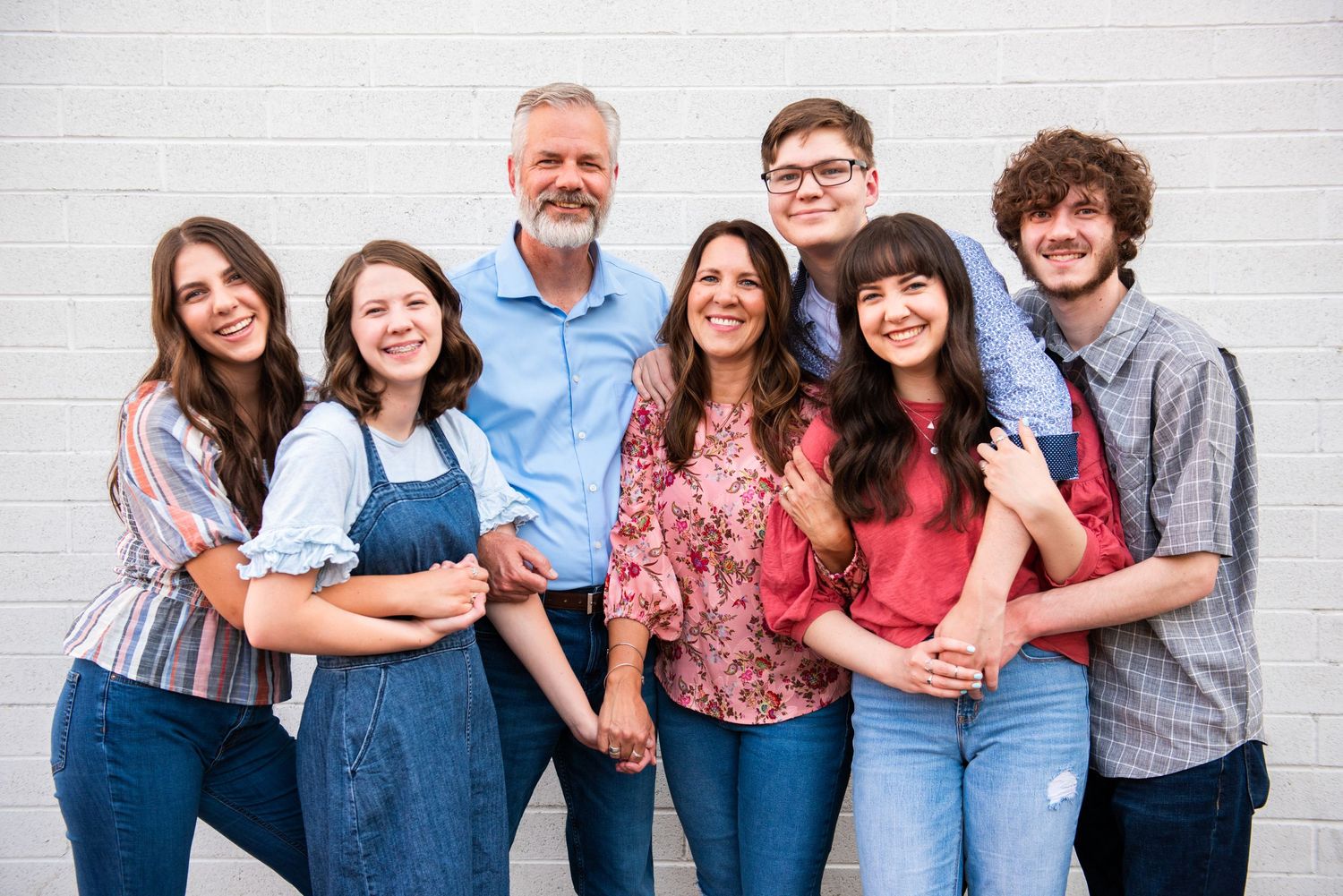A family group poses together against a white brick wall wearing casual clothing.