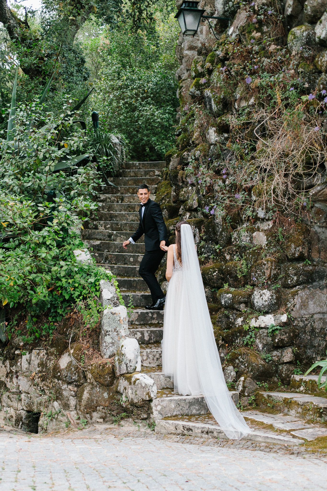 Long wedding veil trails down romantic stone staircase surrounded by greenery.