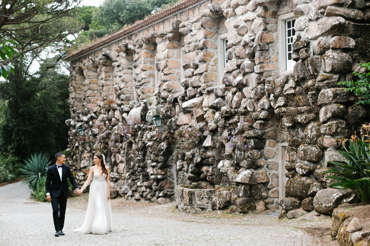 Newlyweds walking along rustic stone wall exterior of historic building.