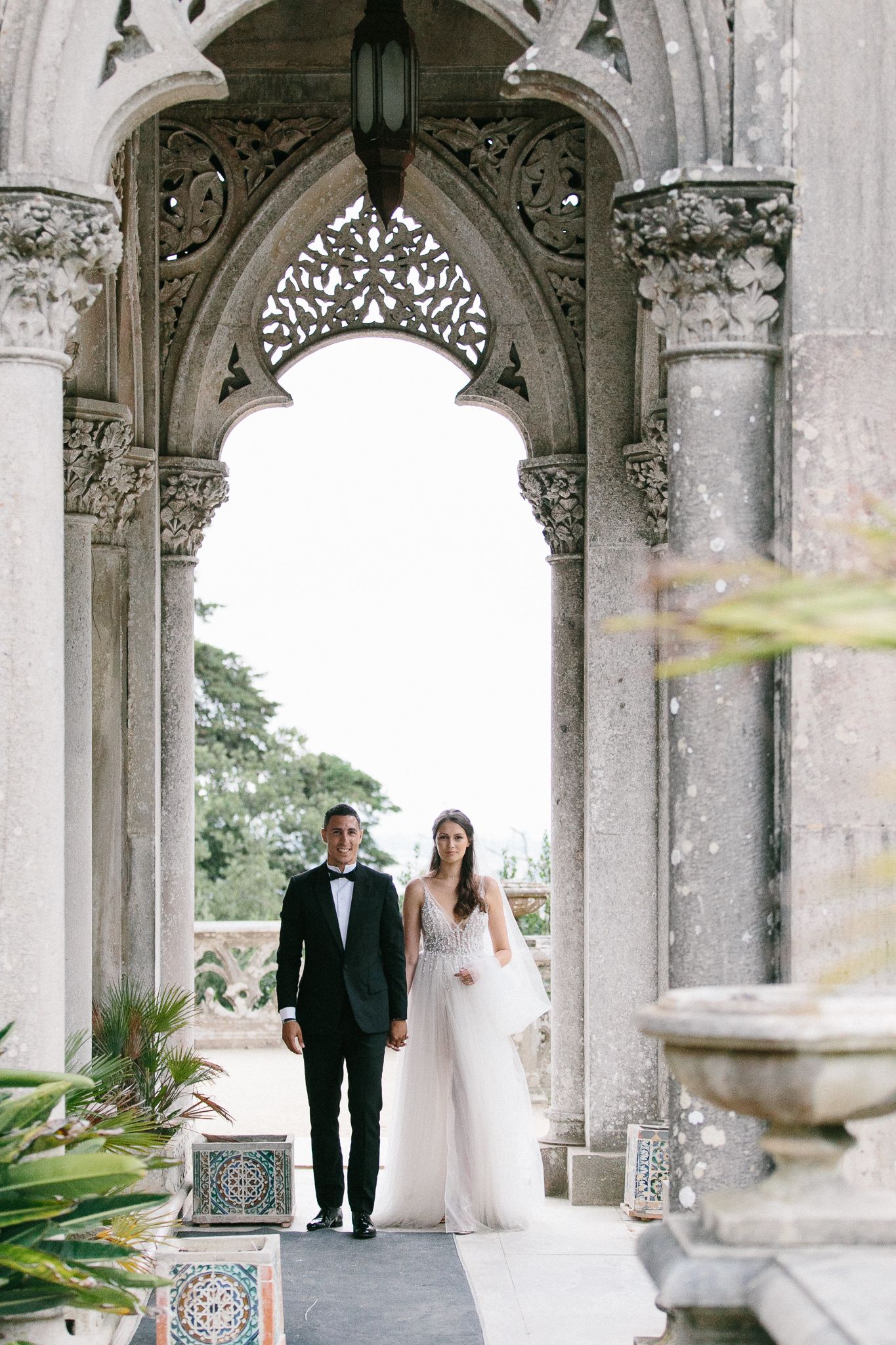 Wedding couple standing under ornate Gothic stone archway with decorative columns.