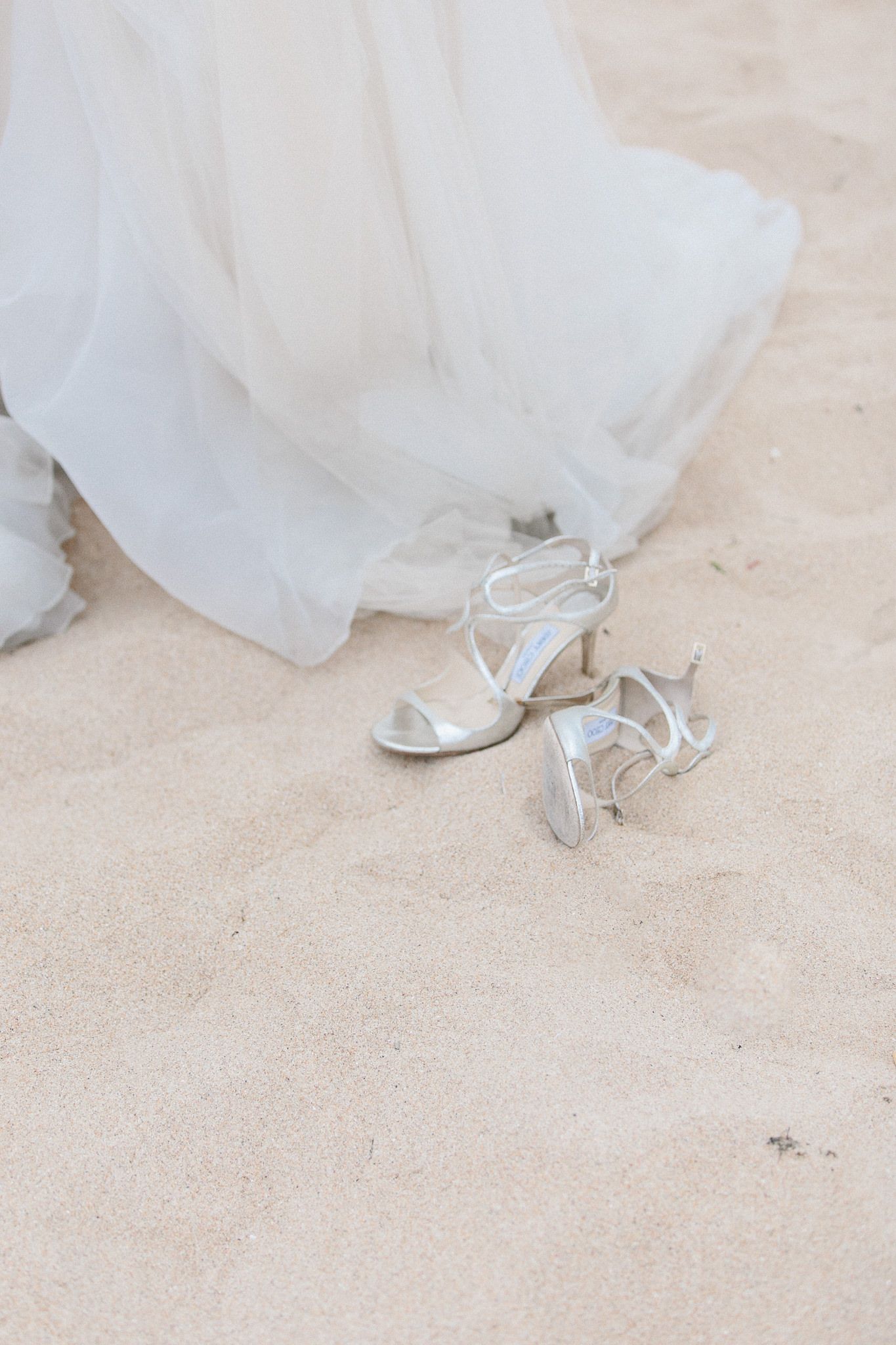 White bridal shoes placed on sand with flowing wedding dress fabric in view.