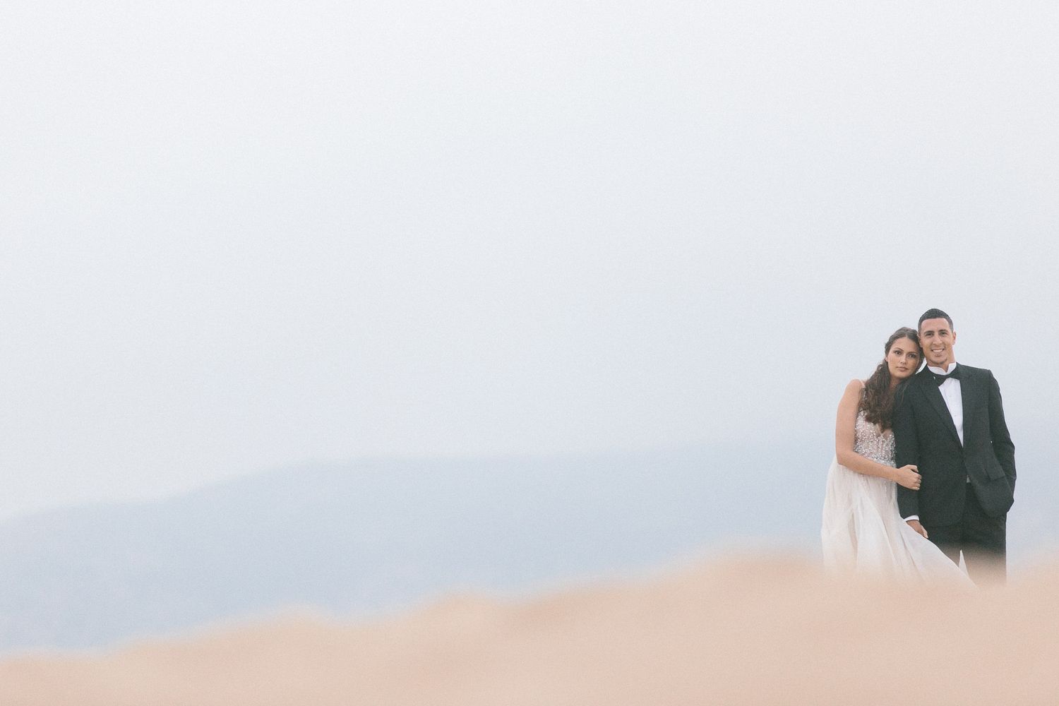 A couple poses together on a sandy dune with misty mountains in the distance.