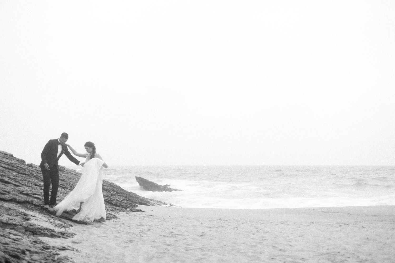 Black and white photo of a couple on rocky beach shore with crashing waves.