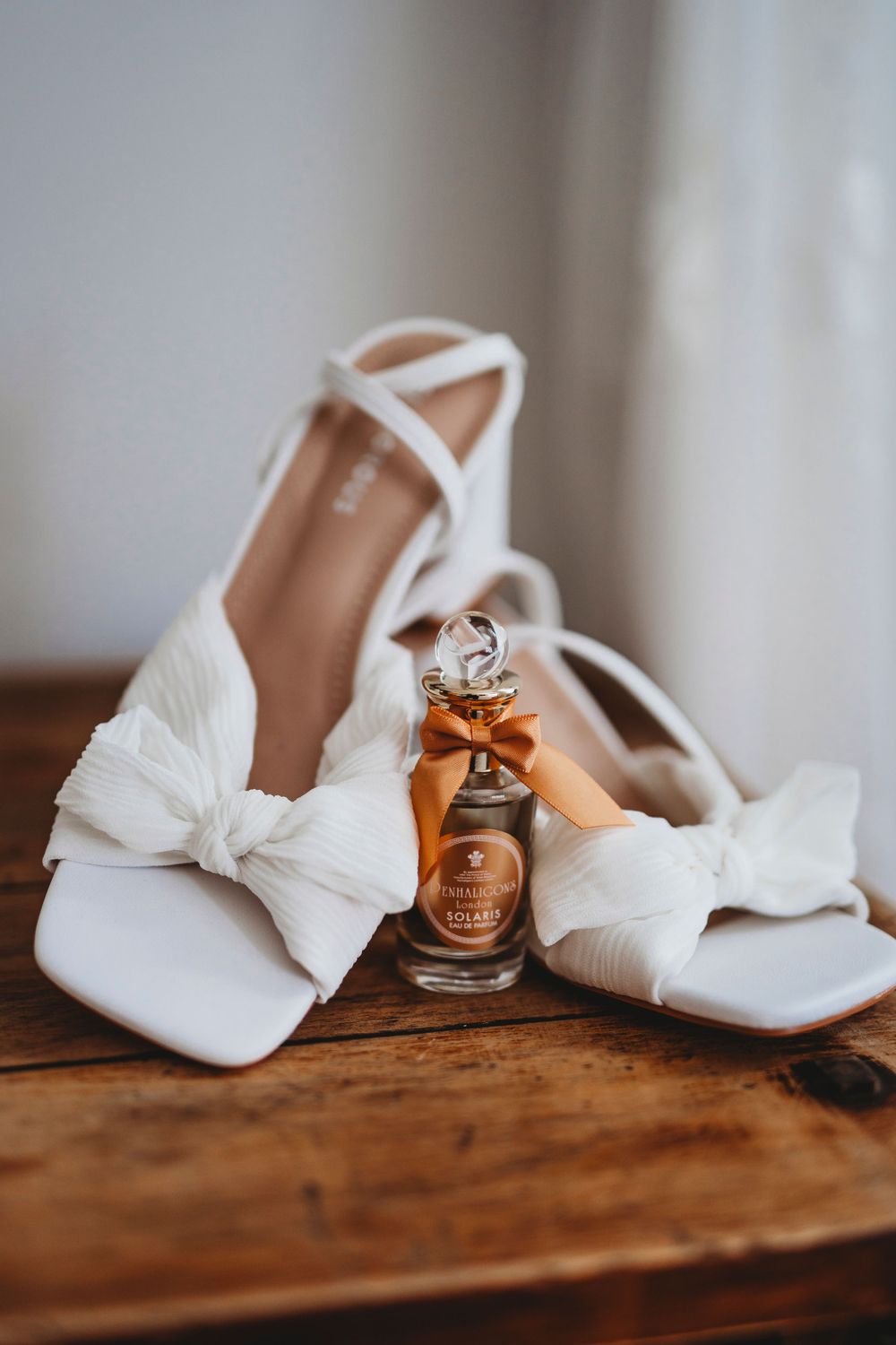 White bridal shoes with bows displayed with a small perfume bottle on wooden surface.