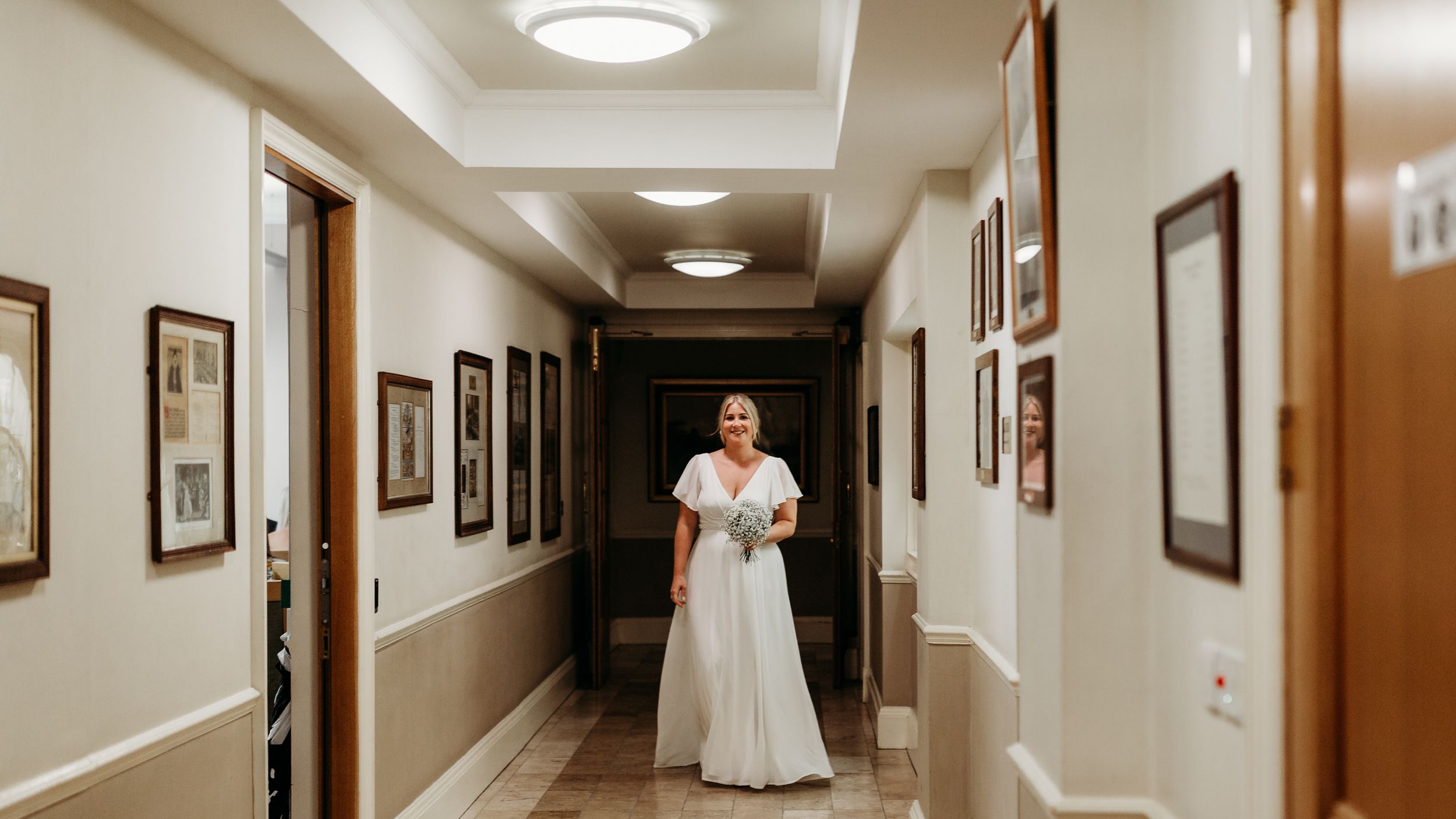 A person in a white wedding dress stands alone in an elegant hallway with framed artwork.