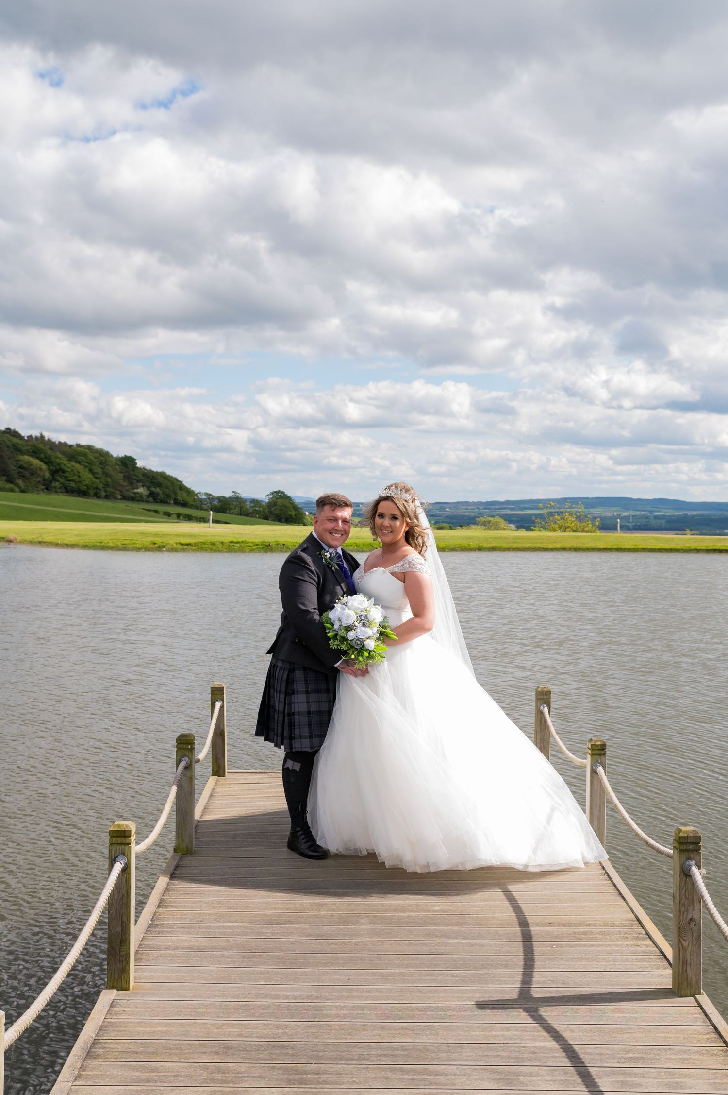 A wedding couple stands on a wooden dock overlooking a lake with cloudy skies above.