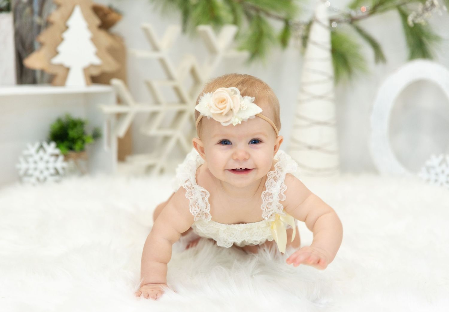 Baby in white dress sitting on soft white blanket with winter holiday decor in background.