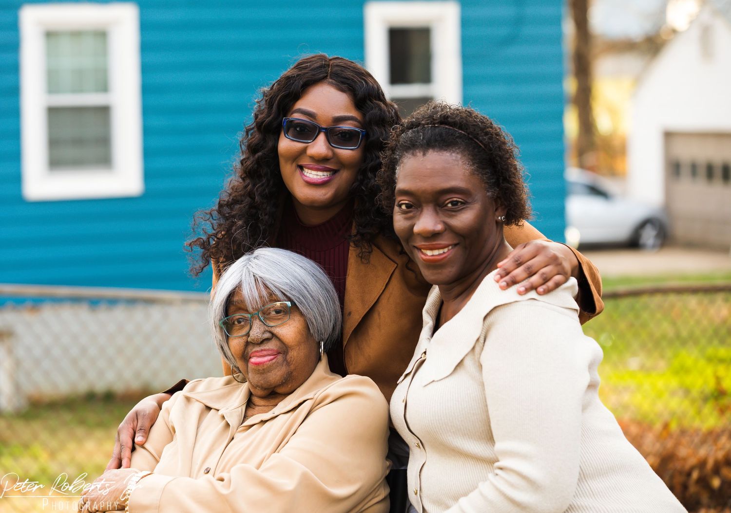 Three generations of women smile together outdoors in front of a blue house and chain-link fence.