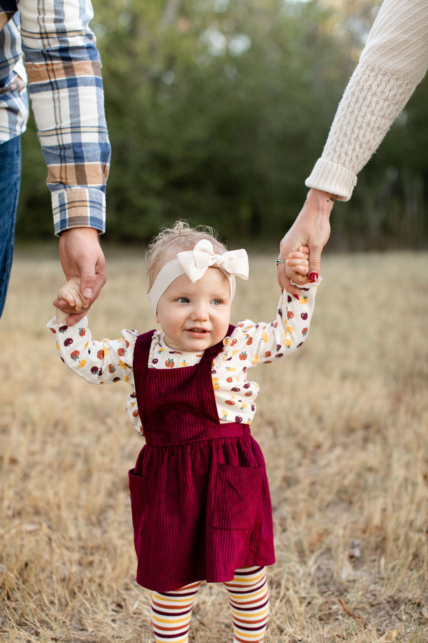 A smiling toddler in burgundy dress holds hands with parents while learning to walk.