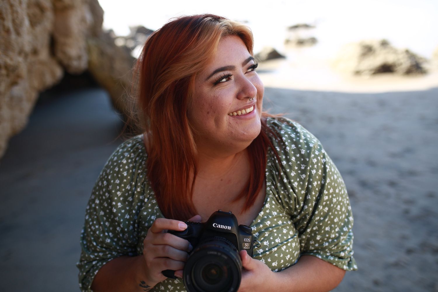 Photographer with red hair smiling while holding camera on beach near cave entrance.