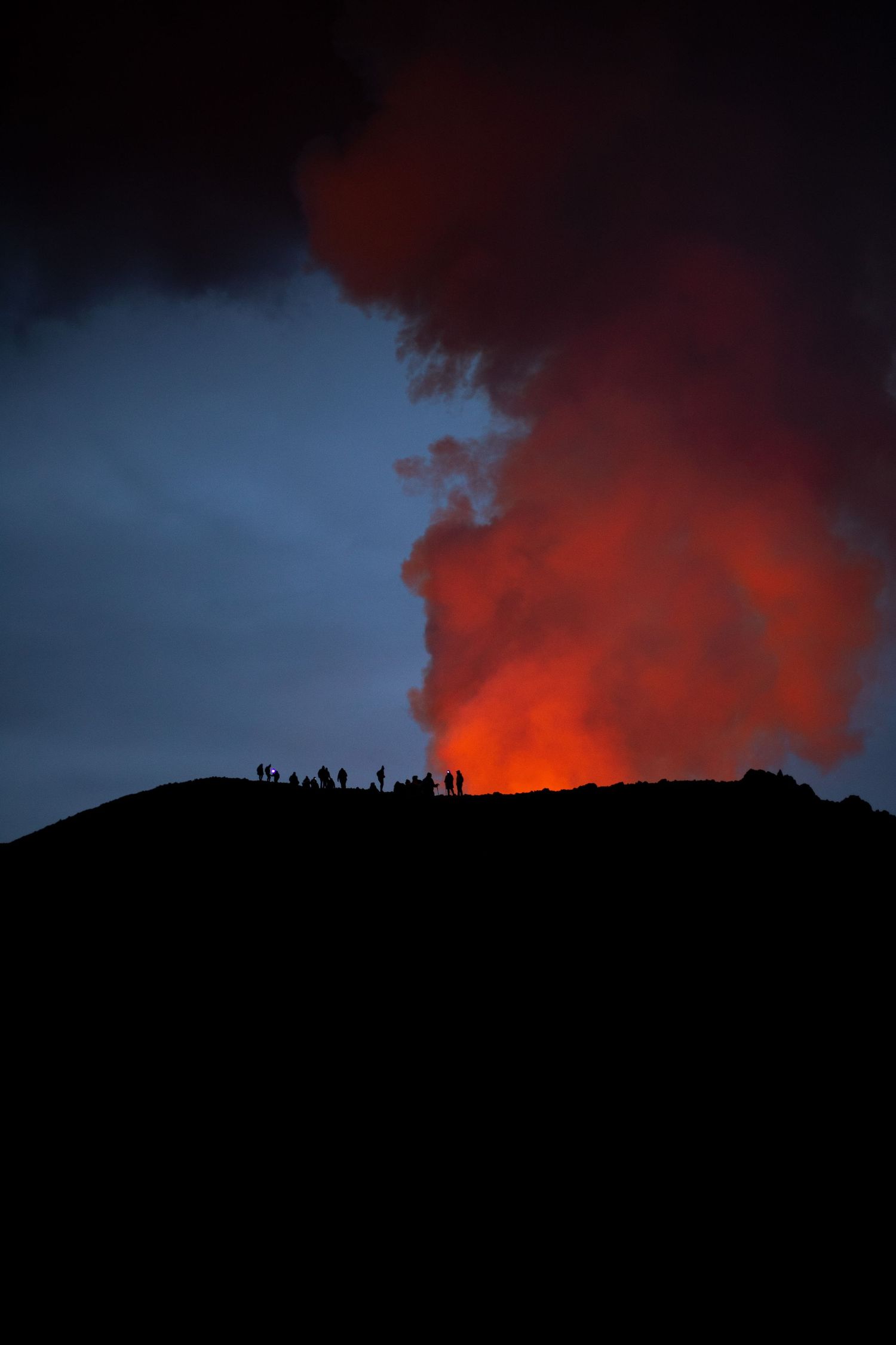 A group of people dressed in hiking equipment are silhouetted against a dark blue sky and a red plume of smoke.