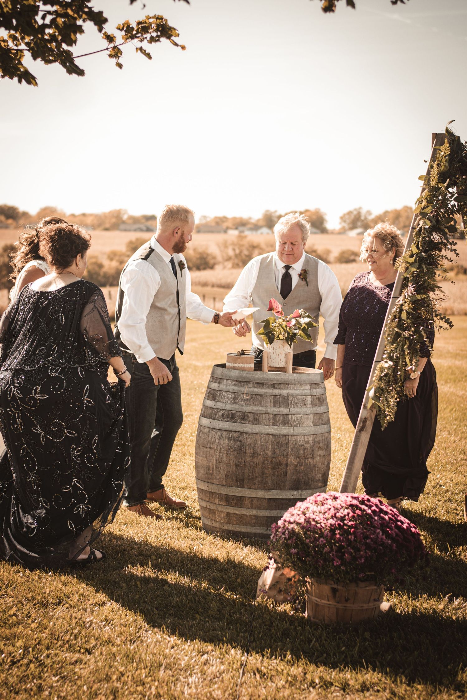 Wedding party surrounding rustic barrel with floral decorations outdoors.