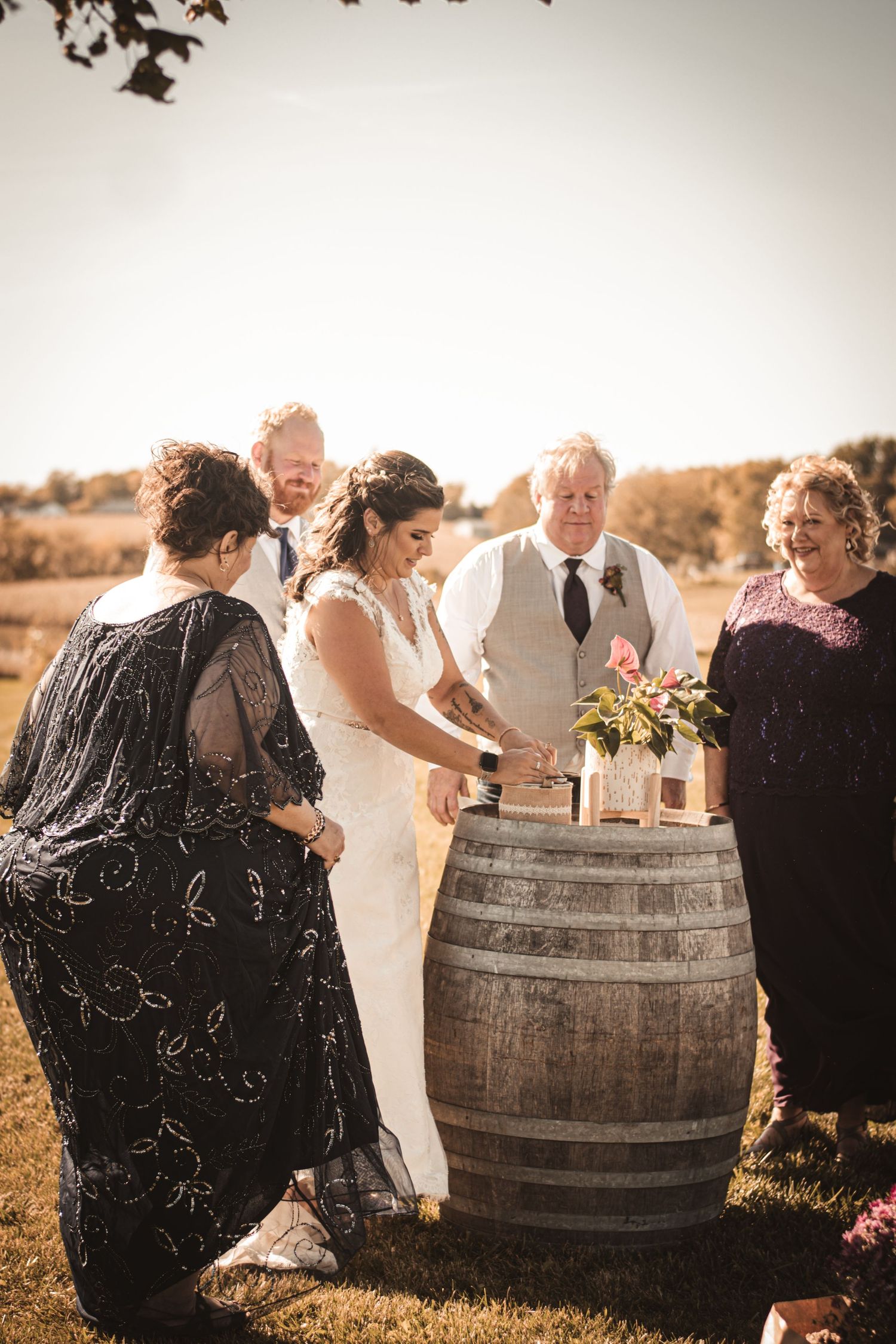 Group gathered around wooden barrel during outdoor wedding ceremony at sunset.
