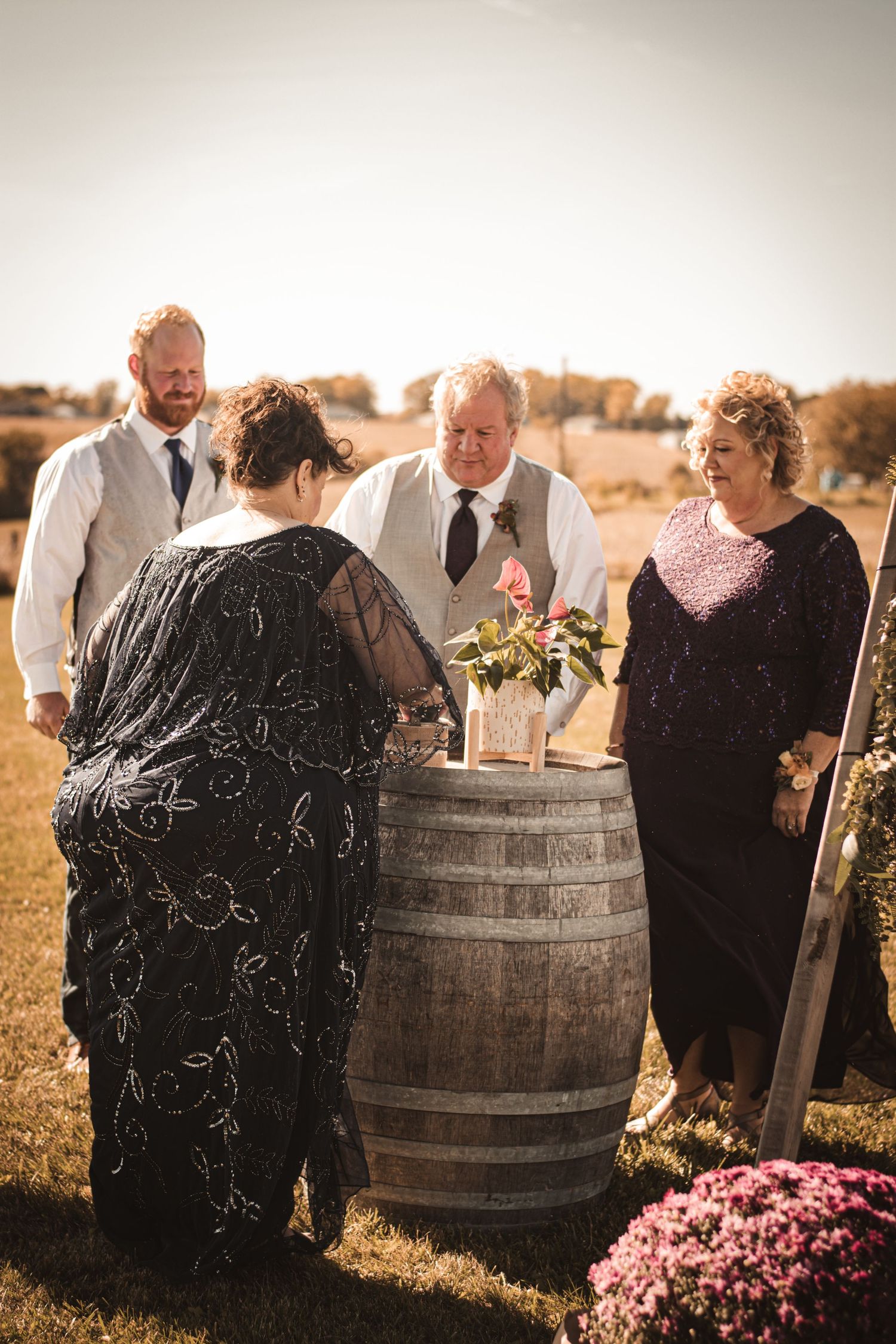 Wedding party members engage in ceremony activities around rustic barrel display outdoors.