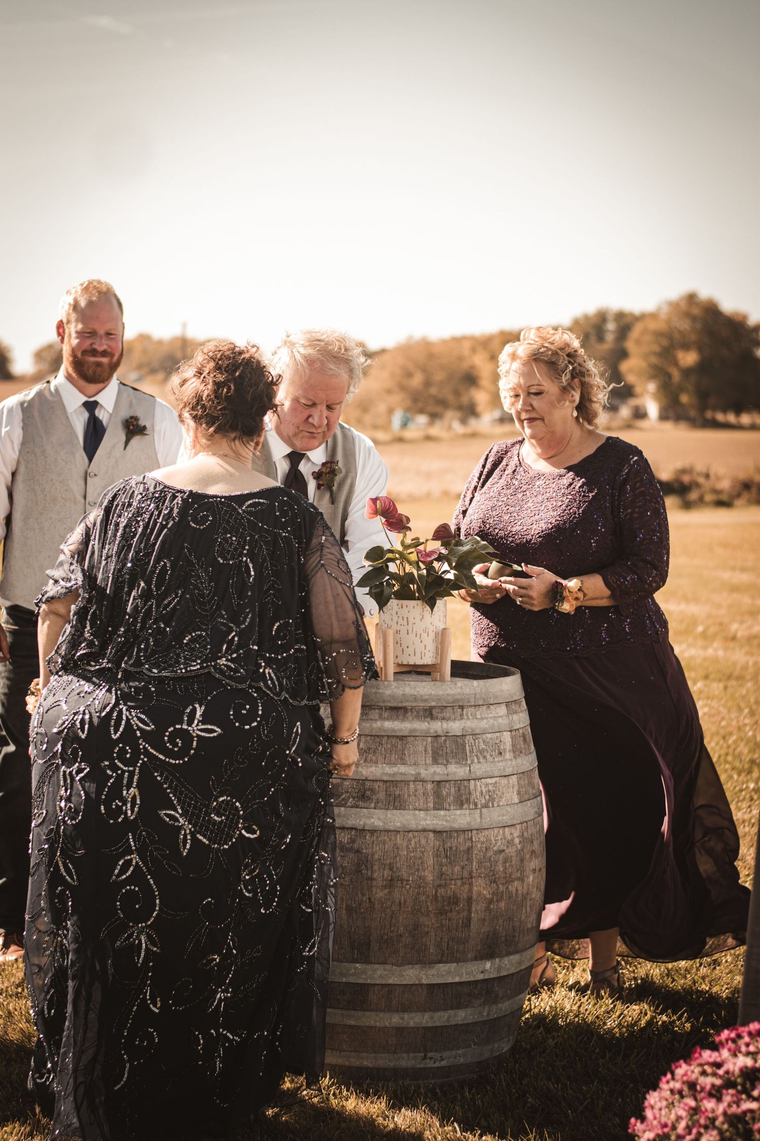 Wedding celebration group gathered around decorative wine barrel in outdoor setting.