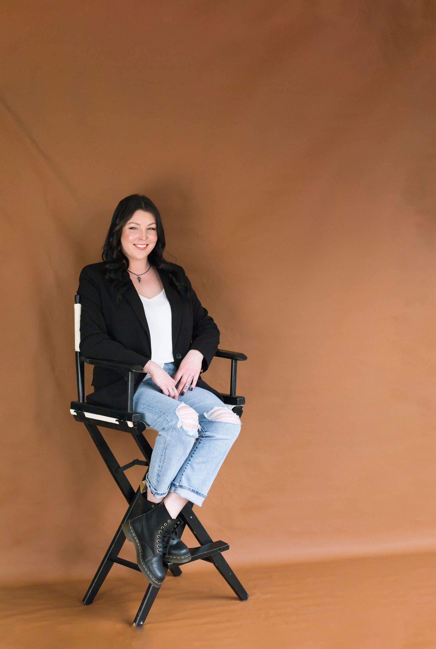 Professional headshot of makeup artist sitting in director's chair against brown backdrop.