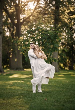 A joyful couple spins together on a grassy lawn surrounded by trees at sunset.