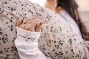 Close-up of intertwined hands with 'I said yes' text on white sleeve against floral pattern.